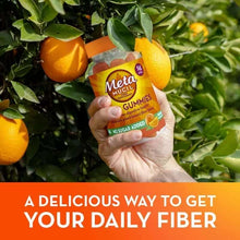 Hand holding Metamucil fiber gummies no sugar added bottle near orange tree with ripe oranges